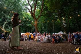 A wedding photographer captures heartfelt speeches under a canopy of trees at Woodland Weddings. The enchanting image shows guests listening intently, illuminated by warm strings of lights.